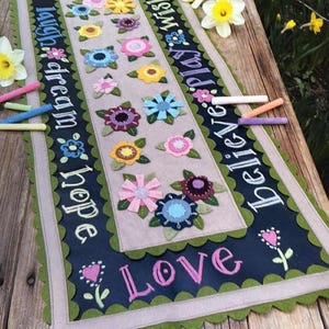 May include: A decorative table runner with a central floral design and inspirational words. The runner features colorful felt flowers in various shapes and colors, with the words "Love, Laugh, Dream, Hope, Believe, Play, Wish" in white script on a dark blue background.