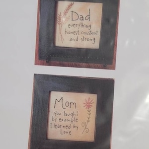 May include: Two framed art pieces with the words "Dad" and "Mom". The "Dad" frame has text that reads "everything honest, constant and strong" with a floral design. The "Mom" frame reads "You taught by example, I learned by Love" with a floral design.