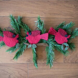 May include: Three red felt cardinal ornaments with black button eyes and brown felt branches. The ornaments are hanging on a green pine garland.
