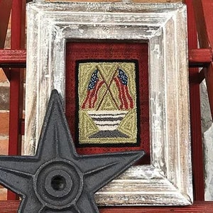 May include: A framed needlepoint artwork featuring two crossed American flags in red, white, and blue, with a white and blue striped bowl in the center. The frame is white with a distressed finish.