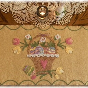 May include: A beige felt table runner with an embroidered design of a basket filled with Easter eggs and flowers. The basket is surrounded by green leaves and pink and yellow flowers.