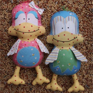 May include: Two handmade plush bird toys. One is pink with blue eyes and a blue bow. The other is blue with white stripes and green accents. Both have yellow beaks and feet.