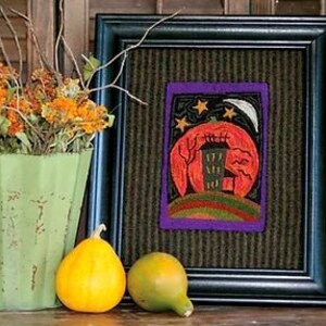 May include: A framed needlepoint artwork depicting a green house with a black door and windows, set against a red and orange sunset with a crescent moon and stars. The background is a dark green and brown landscape.