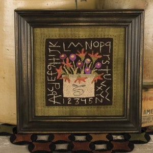 May include: A framed cross-stitch embroidery featuring a bouquet of purple flowers in a white pot. The embroidery is surrounded by a green background with a border of black letters and numbers. The letters spell out "ABC DEF GHI JKL MNO PQR STU VWX YZ" and the numbers spell out "1 2 3 4 5 6 7 8 9 10 11 12".