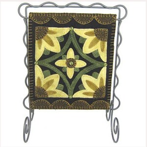 May include: A black, green, and yellow needlepoint wall hanging with a floral design is displayed on a metal stand.