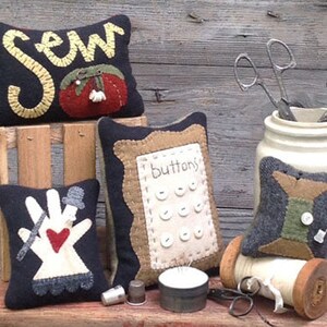 May include: A collection of handmade sewing-themed items. Includes decorative pin cushions with embroidered designs, a jar with scissors, a spool of thread, and a thimble. The cushions feature words and images related to sewing.