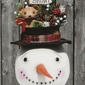May include: A snowman with a black top hat, a red plaid band, and a red bow tie. The snowman has a brown teddy bear wearing a red scarf on top of the hat. The snowman has a carrot nose and black eyes. The snowman is in front of a gray wooden background. The sign on top of the hat says "Let it Snow!"