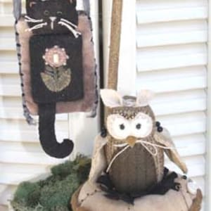 May include: Two handmade felt toys, a black cat with a flower on its chest and a brown owl with a beige nest. The cat is hanging from a brown strap and the owl is sitting on its nest.