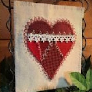May include: A fabric wall hanging featuring a large red heart with a white lace accent. The heart is stitched onto a cream-colored background. The heart's design incorporates a patchwork pattern and a scalloped edge.