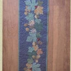 May include: A blue quilted table runner with a design of falling leaves, acorns, and berries in shades of green, brown, and red.