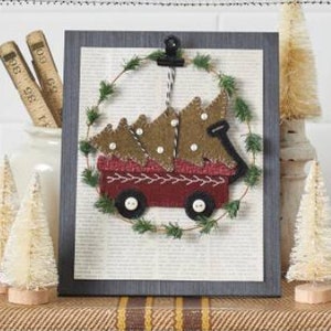 May include: A framed felt Christmas ornament depicting a red wagon carrying a stack of three green and brown felt Christmas trees. The wagon is decorated with white felt dots and a black felt trim. The ornament is surrounded by a green felt wreath.