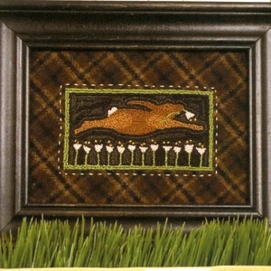 May include: Framed artwork featuring a brown rabbit in mid-leap, set against a dark plaid background. The rabbit is surrounded by a green border with white flower-like shapes at the bottom. The frame is dark brown.