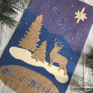 May include: A felt Christmas stocking with a blue and purple background. The stocking features a deer, two trees, and a star. The text "All is Calm... All is Bright" is embroidered on the stocking.