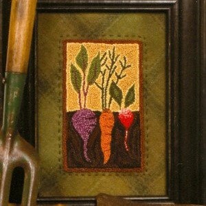May include: Framed artwork featuring a needlepoint design of root vegetables: a purple beet, an orange carrot, and a red radish. The artwork is set against a textured green background within a black frame. A gardening tool is visible in the foreground.