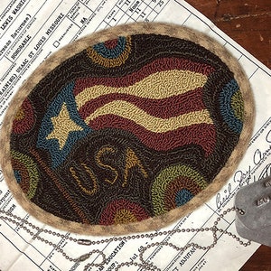 May include: A close-up of a needlepoint design featuring an American flag with a star and the word "USA" in a circular frame. The design is on a brown background with a beige border. The design is surrounded by a silver chain and two silver dog tags.