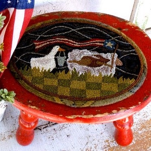 May include: A small, distressed red wooden stool with a round top. The top features a hand-stitched, oval rug with a patriotic design: a goose, sheep, and American flag. The rug has a black background and a checkered green and brown border.