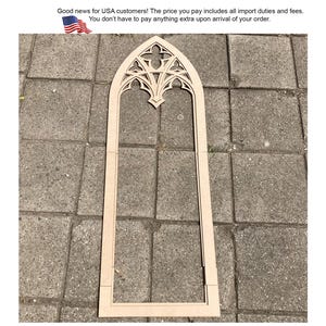 May include: A tall, light-coloured wooden frame shaped like a pointed gothic arch window. The top section features intricate, symmetrical cut-out tracery patterns, while the lower portion consists of straight vertical sides and a flat base.