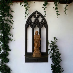 Op de afbeelding: Een houten beeld van de Maagd Maria die een baby Jezus vasthoudt, tentoongesteld in een zwart gotisch raamwerk met een plank. Het frame is gemonteerd op een witte muur met groene planten aan beide kanten.