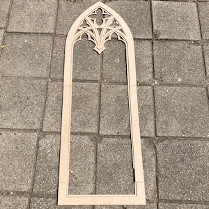 May include: A light brown, unfinished wooden gothic arch window frame. The top section features an ornate, cut-out design, while the frame is rectangular. The frame is made of multiple pieces of wood.