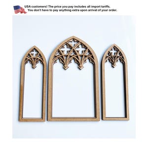May include: Three light brown wooden window frames with pointed gothic arches. The frames feature intricate cutout tracery patterns at the top, including quatrefoil shapes. The set consists of one wider central frame and two narrower side frames.