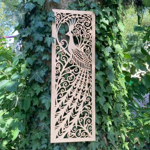 Peut inclure: Panneau rectangulaire en bois découpé au laser représentant un paon. Le paon est représenté avec des détails complexes, entouré de motifs tourbillonnants et d'éléments floraux. Le panneau est de couleur marron clair et est présenté sur un fond de feuillage vert.