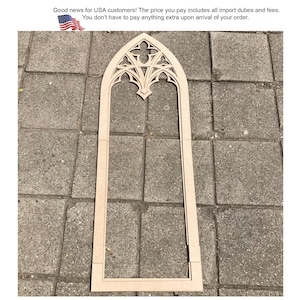 May include: A tall, light-colored wooden frame shaped like a pointed gothic arch window. The top section features intricate, symmetrical cut-out tracery patterns, while the lower portion consists of straight vertical sides and a flat base.
