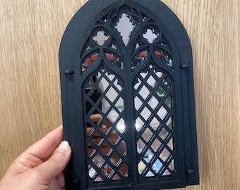 May include: Black gothic-style mirror with an arched top and intricate cut-out details. The mirror features a diamond-patterned design and is held by a hand. The mirror is made of black material and reflects the surrounding environment.