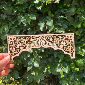 Op de afbeelding: Een lichtbruin, lasergesneden houten decoratief paneel met een ingewikkeld bloemen- en arabeskpatroon. Het paneel heeft een geschulpte onderrand. De achtergrond is een dichte groene haag.