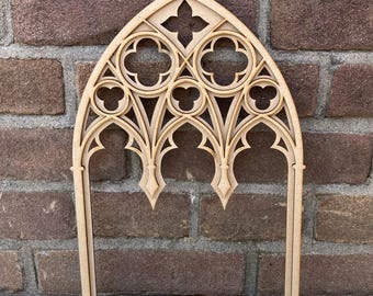 Puede incluir: Un marco de ventana de arco gótico de madera marrón claro con intrincados detalles recortados. El marco presenta un diseño de cuatro hojas en la parte superior y formas de arco apuntadas. El marco está sobre una pared de ladrillos.