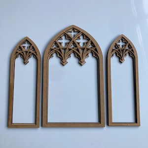 May include: Three wooden gothic arch frames in different sizes. The frames are light brown and have detailed cut-out patterns at the top. The arches are empty, giving a window-like look. The frames are arranged on a white surface.