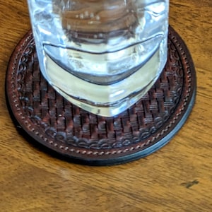 May include: A brown leather coaster with a woven pattern and a decorative border. The coaster is designed to protect surfaces from heat and moisture.