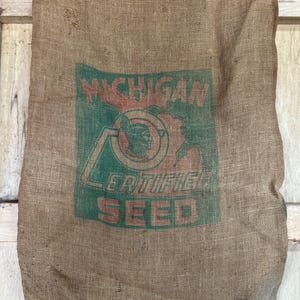 May include: A large, tan burlap sack with a green and red graphic that reads "MICHIGAN CERTIFIED SEED." The sack has a worn, vintage appearance, with visible wear and tear. The graphic features a stylized image of a person.