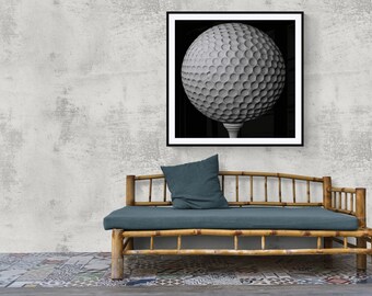 Op de afbeelding: Een zwart-witfoto van een golfbal op een tee, ingelijst in een zwart frame. De foto hangt aan een grijze muur boven een bamboe bank met een blauw kussen.