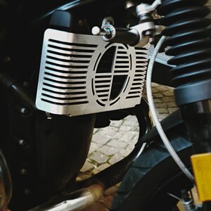 May include: A silver motorcycle radiator guard with a horizontal striped design and a central oval cutout featuring a stylized emblem. The guard is mounted on a black motorcycle, with visible suspension components.