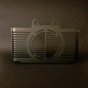 May include: A black metal component with a rectangular body and a circular design on the front. The component has horizontal lines and two small, rounded tabs at the top. The design is symmetrical and geometric.