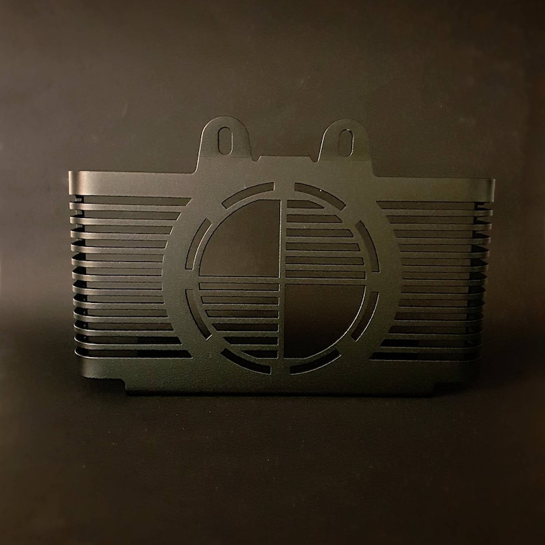 May include: A black metal component with a rectangular body and a circular design on the front. The component has horizontal lines and two small, rounded tabs at the top. The design is symmetrical and geometric.