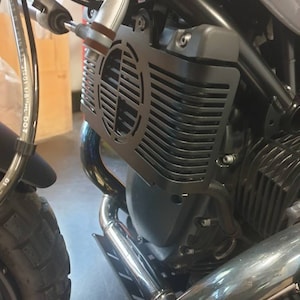 May include: Close-up of a motorcycle engine with a black metal radiator guard. The guard has a circular, slotted design. Chrome exhaust pipes and a black engine block are also visible.