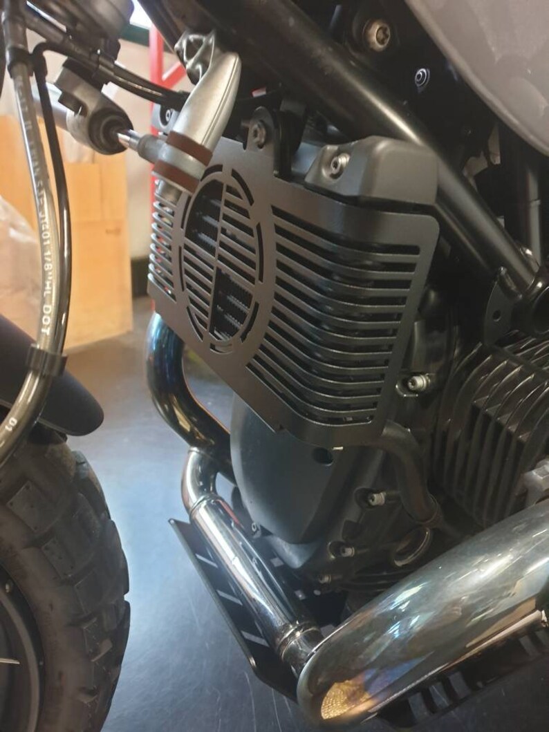 May include: Close-up of a motorcycle engine with a black metal radiator guard. The guard has a circular, slotted design. Chrome exhaust pipes and a black engine block are also visible.