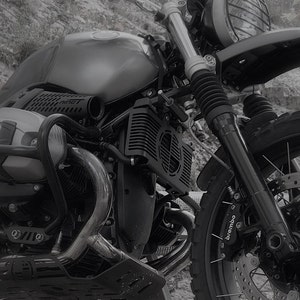 May include: A grayscale image of a motorcycle, showcasing its front section. The motorcycle features a round headlight, a fuel tank, and a prominent engine with a radiator. The front suspension and a Brembo brake caliper are also visible.