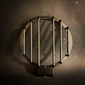 May include: A dark gray metal headlight guard with a circular frame and vertical bars. The guard has a mounting bracket at the bottom and is designed to protect a vehicle's headlight.