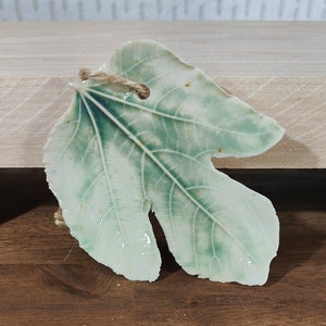 May include: A ceramic fig leaf ornament with a green and white glaze. The leaf has a rustic, hand-crafted look and is strung with twine for hanging.