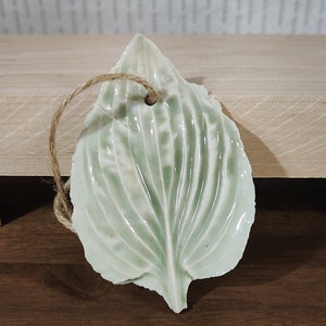 May include: A ceramic leaf ornament with a light green glaze and a textured surface. The leaf has a hole at the top for hanging.
