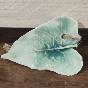 May include: A ceramic leaf-shaped ornament with a light green glaze and detailed veining. A hole is at the top with a natural twine string attached. The leaf rests on a dark brown wooden surface, with a light wood background.