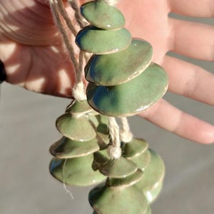 Puede incluir: Adorno de cerámica artesanal con formas verdes redondeadas apiladas, unidas con hilo natural. El acabado brillante de la cerámica refleja la luz, resaltando las diferentes tonalidades de verde. Diseñado para colgar.