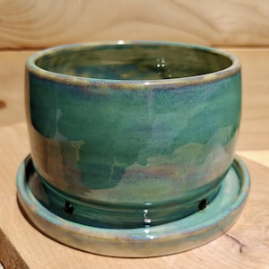 May include: A handcrafted ceramic planter with a matching saucer. The planter is a vibrant teal color with a glossy finish and a slightly textured surface. The rim and saucer edges have a subtle brown and blue glaze. The planter is designed for indoor or outdoor use.
