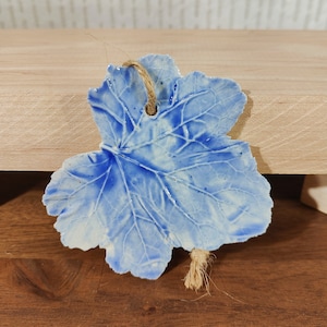 May include: A ceramic leaf ornament with a blue and white glaze. The leaf has detailed veining and a hole at the top for hanging with a natural twine. The ornament is a decorative item.