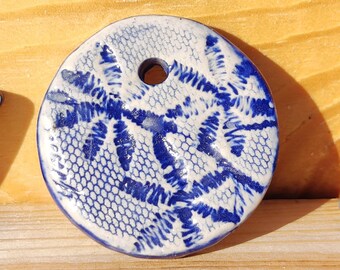 May include: A circular ceramic pendant with a blue and white lace design. The pendant has a hole for threading. The design features a detailed pattern in blue against a white backdrop. The pendant is likely handmade, with a unique, intricate pattern.