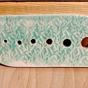 Peut inclure: Une pièce rectangulaire en céramique vert d'eau avec un motif de feuilles et six trous de tailles variables. La pièce en céramique est posée sur une surface en bois.
