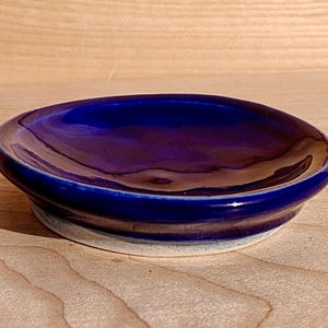 Könnte beinhalten: Eine kleine, runde Keramikschale mit tiefblauer Glasur. Die Schale hat einen leicht erhöhten Rand und einen weißen Sockel. Die glänzende Oberfläche reflektiert das Licht. Die Schale steht auf einer Holzoberfläche. Durchmesser ca. 10 cm.