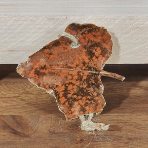 May include: A ceramic leaf-shaped ornament with an orange and brown pattern. It has a naturalistic vein design and a jute string for hanging. The ornament is on a wooden surface.